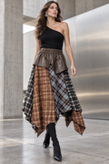 A model walks in a modern concrete and stainless steel corridor wearing a unique patchwork skirt with brown plaid, black-and-white plaid, and brown faux-leather panels, paired with a black one-shoulder top and black boots.