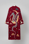 A plush burgundy velvet robe with a large embroidered dragon design on the back, featuring a crescent moon, clouds, and small decorative motifs in teal, gold, and cream, hanging in a boutique.